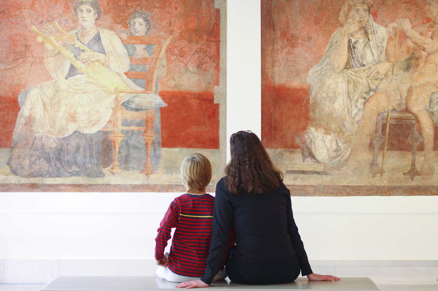 A woman and a young boy sitting together on a bench looking up at two Roman frescoes